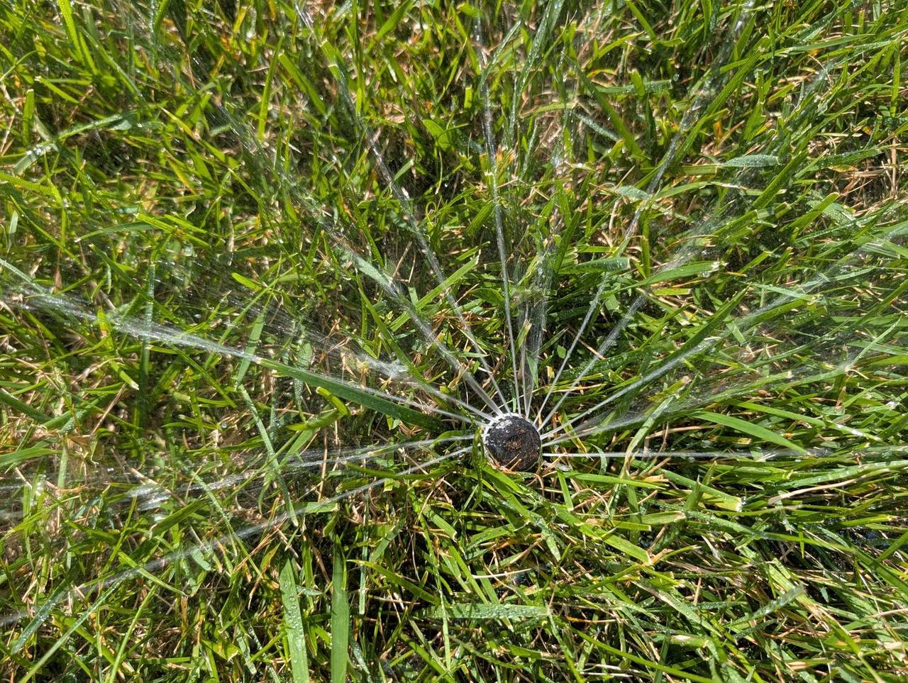 Overhead close-up of a rotary sprinkler head throwing radial jets of water across a green lawn from a professionally installed irrigation system
