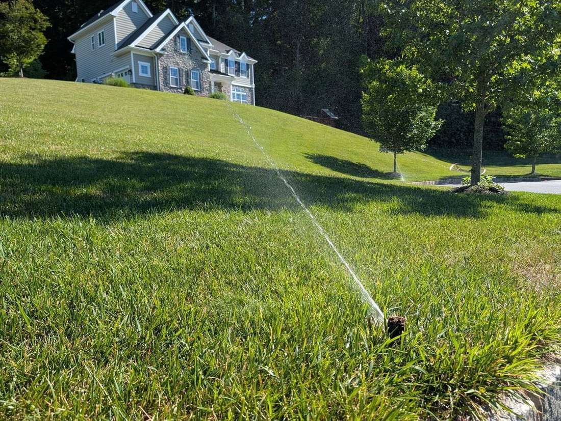 A residential irrigation sprinkler throwing a long stream of water across a manicured sloped lawn toward a stone colonial home in Delaware County, PA